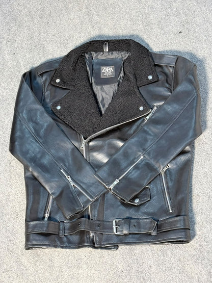 A black leather motorcycle jacket with a black fleece collar, laid flat on a gray textured surface. It features an asymmetrical zipper, zippered chest pockets, and a matching leather belt with a silver buckle.