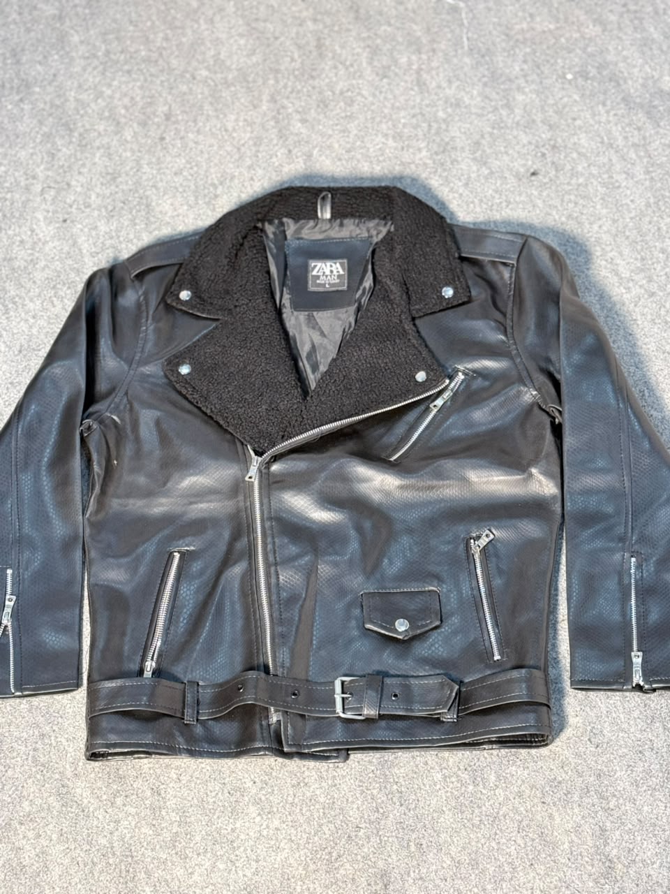 A black faux-leather biker jacket with a dark gray fleece collar, laid flat on a gray textured surface. The jacket features a zippered front, zippered pockets, and a matching belt.