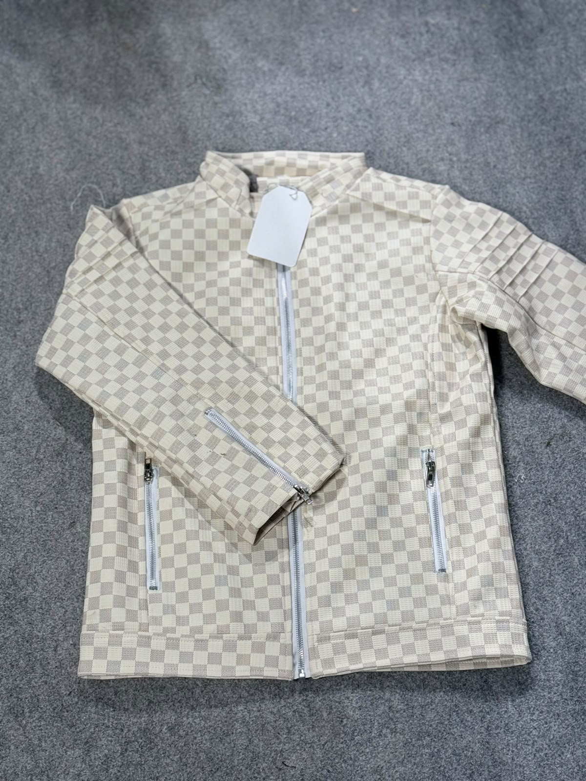 A beige and white checkerboard patterned zip-up jacket with two front zip pockets, laid flat on a gray textured surface.