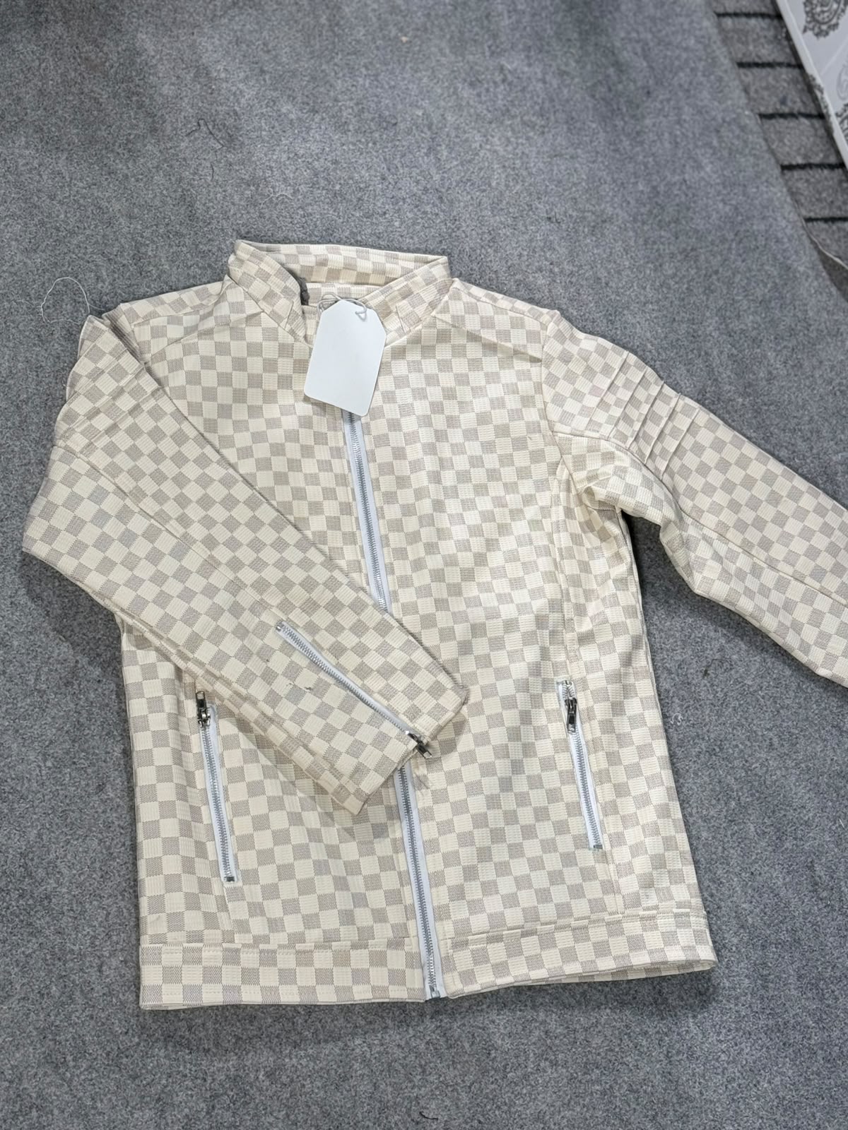 A beige and off-white checkerboard patterned zip-up jacket laid flat on a gray textured surface, with a white hangtag attached.