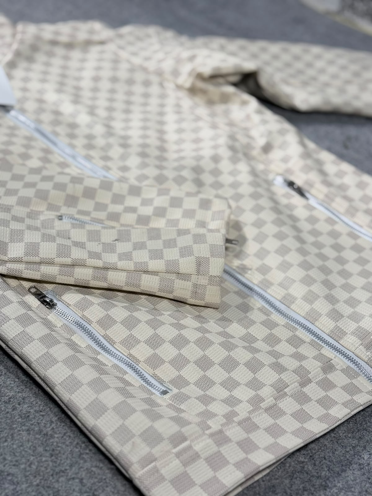A beige and gray checkerboard patterned jacket with silver zippers, laid flat on a gray textured surface.