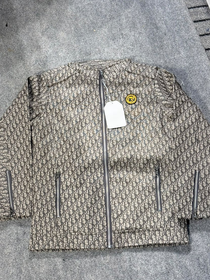 A quilted puffer jacket featuring an all-over beige and brown monogram pattern, a central silver zipper, and zippered pockets, laid flat on a gray textured surface.