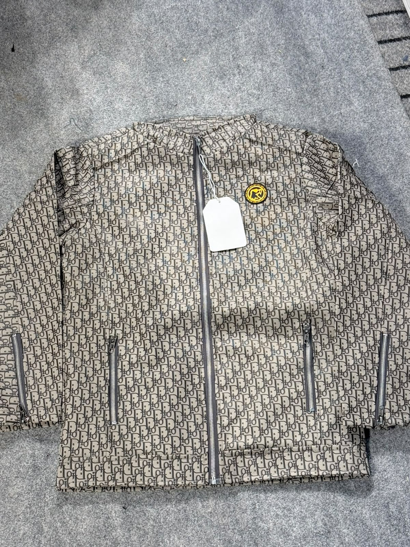 A quilted puffer jacket featuring an all-over beige and brown monogram pattern, a central silver zipper, and zippered pockets, laid flat on a gray textured surface.