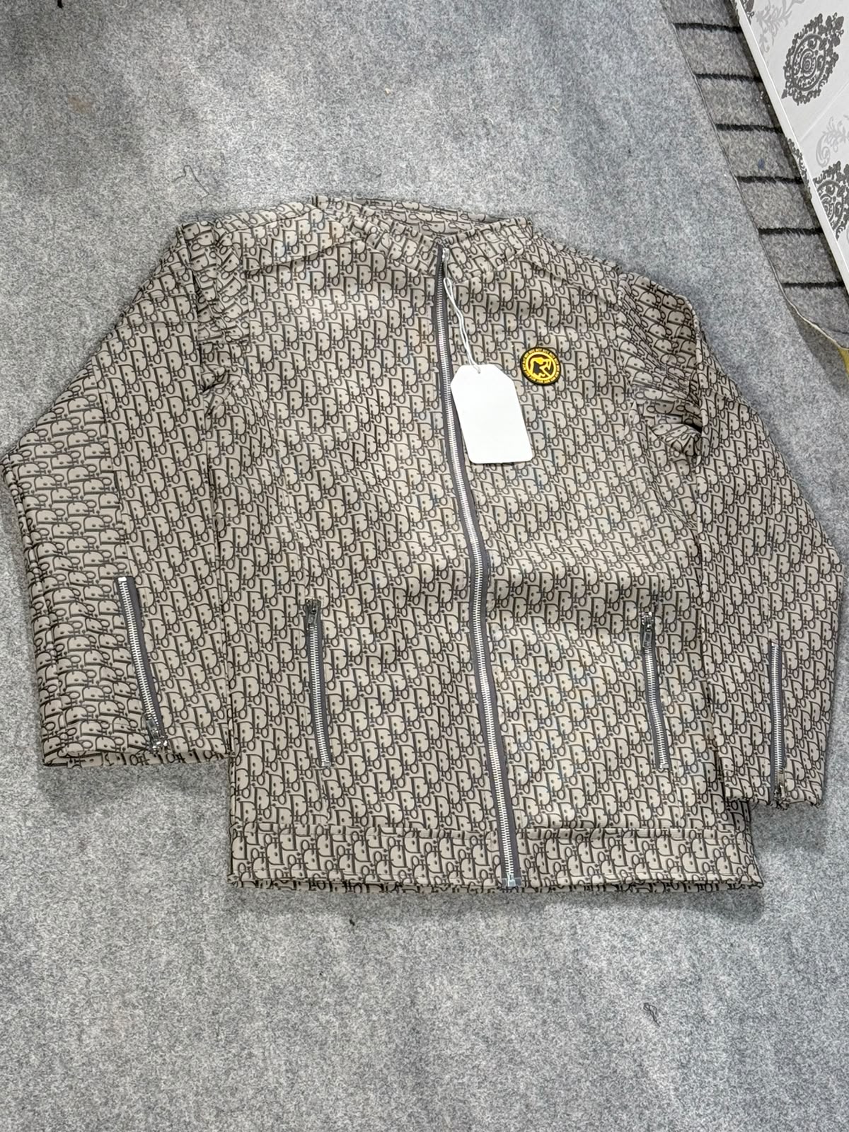 A beige and brown monogram patterned zip-up jacket laid flat on a gray textured surface, featuring a white hangtag and a small yellow smiley face patch on the chest.
