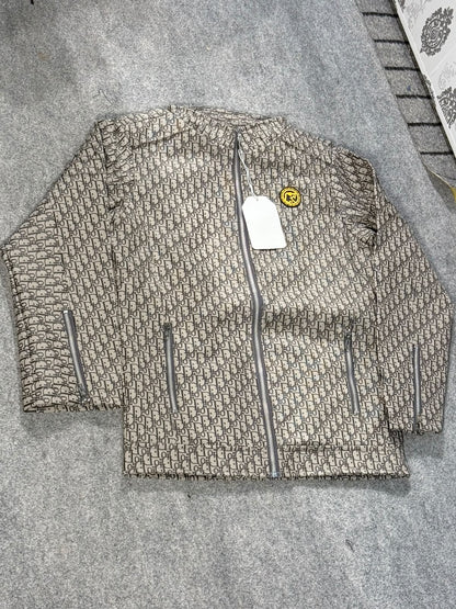 A beige and brown monogram patterned zip-up jacket laid flat on a gray textured surface, featuring a white hangtag and a small yellow smiley face patch on the chest.