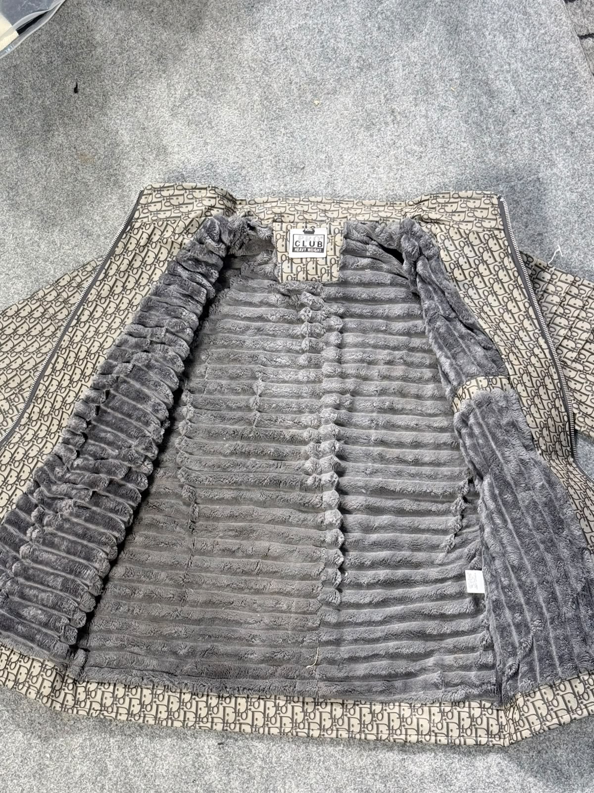 A luxury faux fur jacket with a beige and brown monogram patterned border, laid flat on a gray carpet. The brand 'Dior' is visible on the inner neck label.