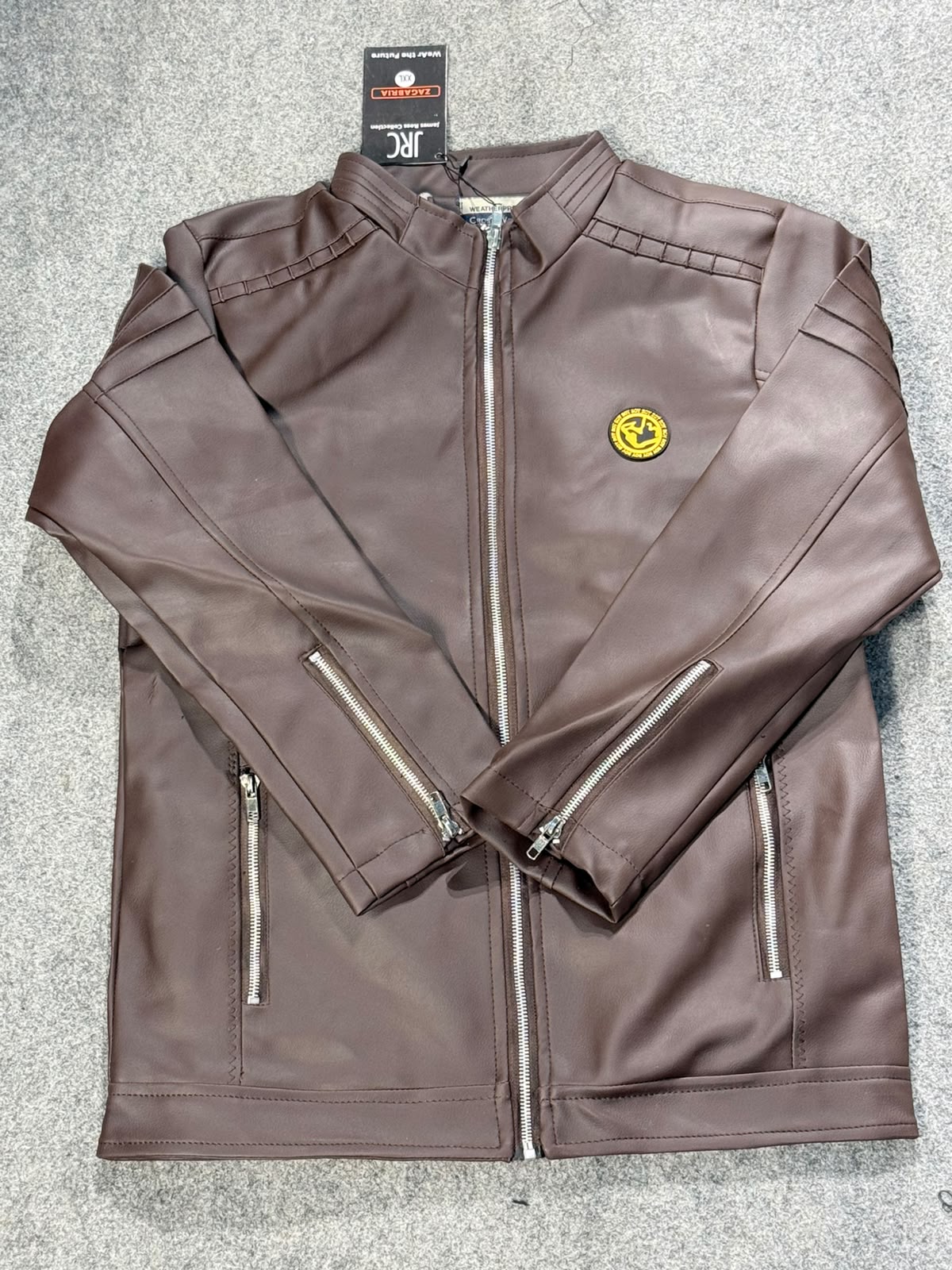 A brown faux leather jacket with silver zippers and a small yellow circular patch on the chest, laid flat on a gray textured surface with a brand tag attached.