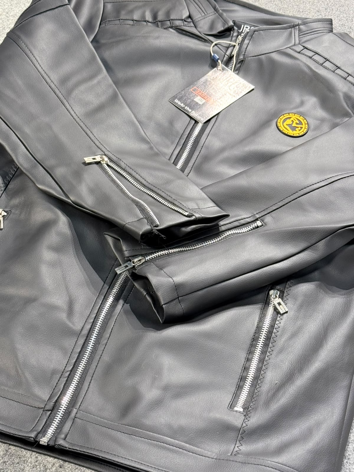 A black faux-leather jacket with silver zippers and a circular yellow logo patch on the chest, shown with its retail hangtag on a gray carpeted floor.