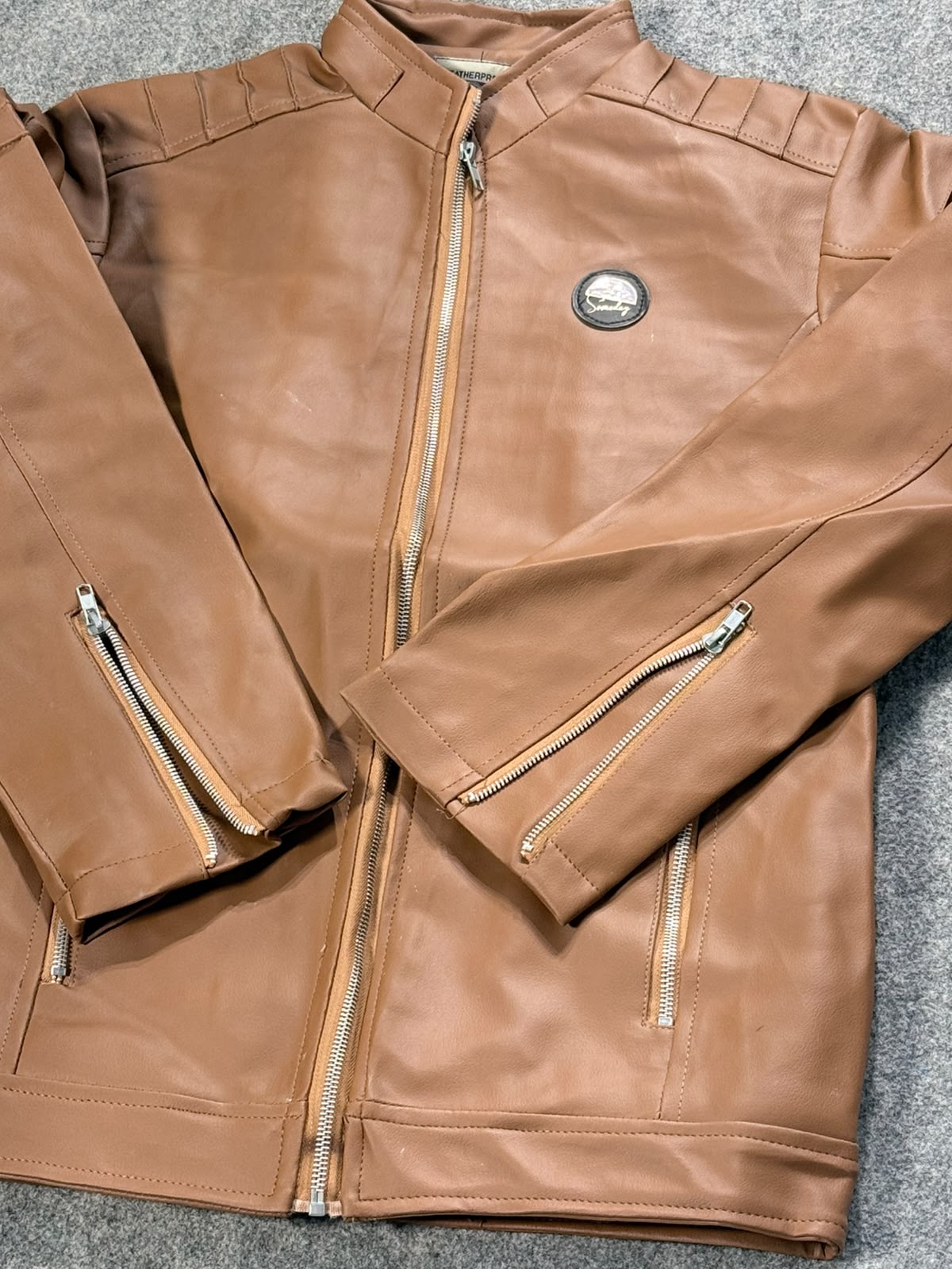 A tan leather motorcycle jacket with silver zippers and a circular metal emblem on the chest, laid flat on a gray textured surface.