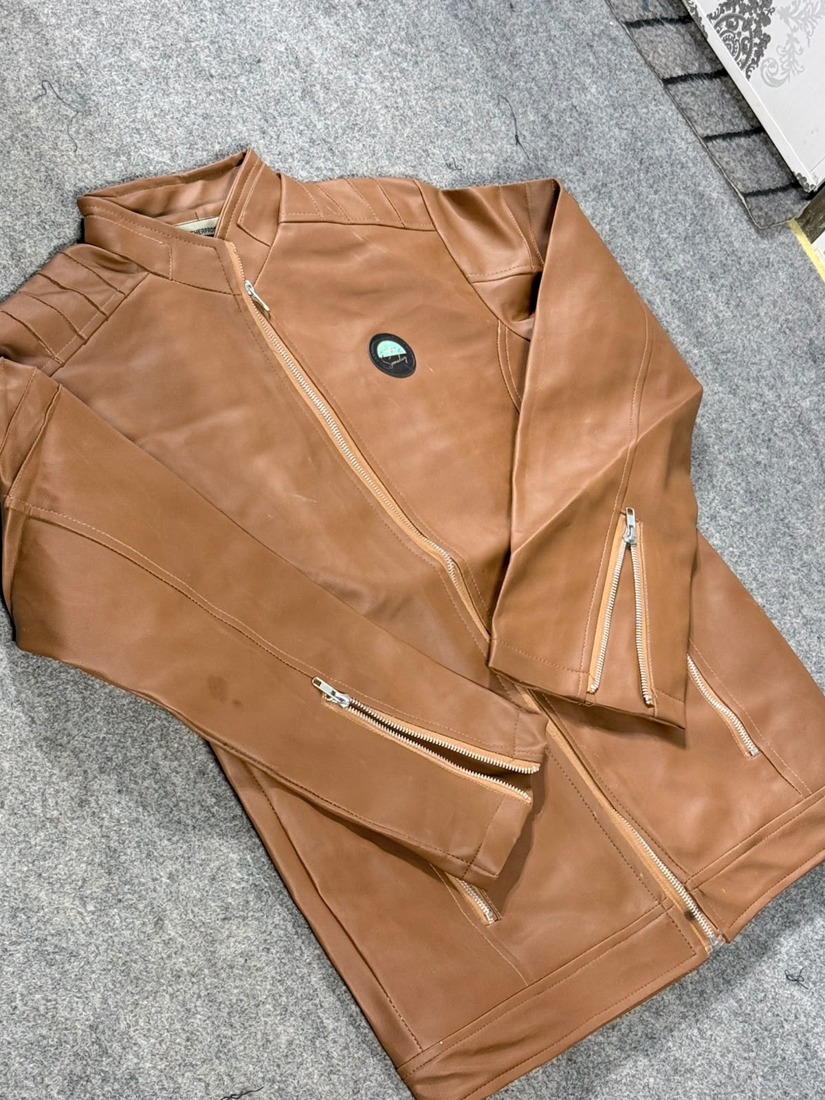 A tan faux-leather motorcycle jacket with multiple zippered pockets, laid flat on a gray textured carpet.