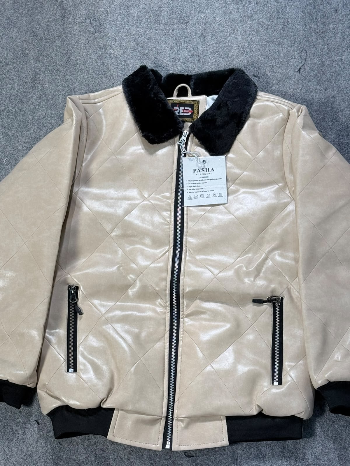 A quilted beige faux-leather bomber jacket with a black faux-fur collar, black ribbed cuffs and hem, and black zipper details, shown flat on a gray textured background. A hangtag from 'FENIA' is visible.