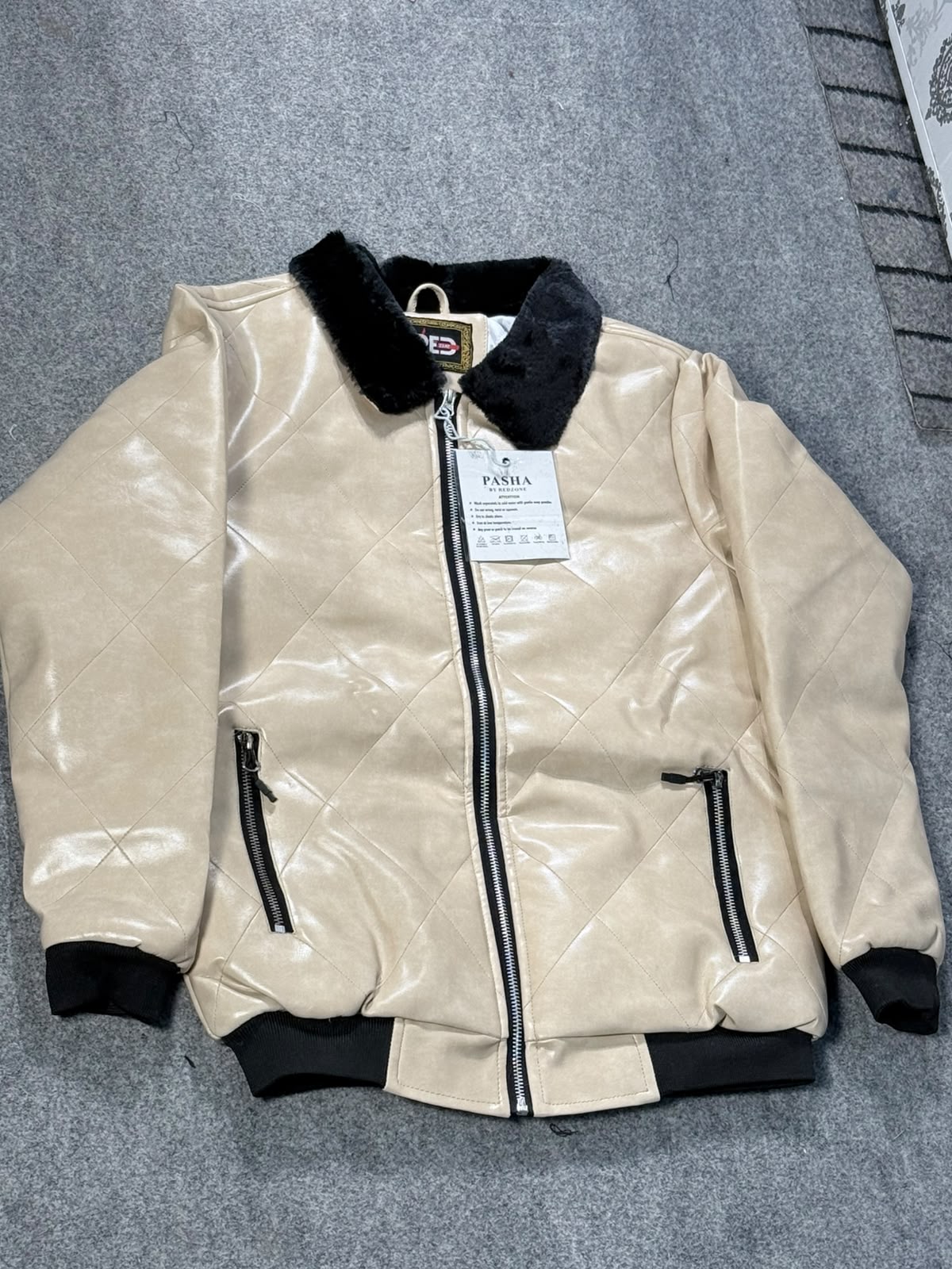 A beige faux-leather bomber jacket with black trim, a black collar, and black zipper details, laid flat on a gray textured surface. A white brand tag for 'FASHION' is attached to the front zipper.