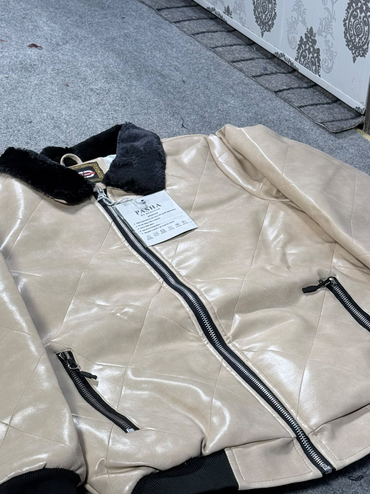 A beige faux-leather bomber jacket with a black faux-fur collar, laid flat on a gray carpeted floor. The jacket features a front zipper, diagonal quilting, and zippered hand pockets, with a visible 'PARIA' brand tag.