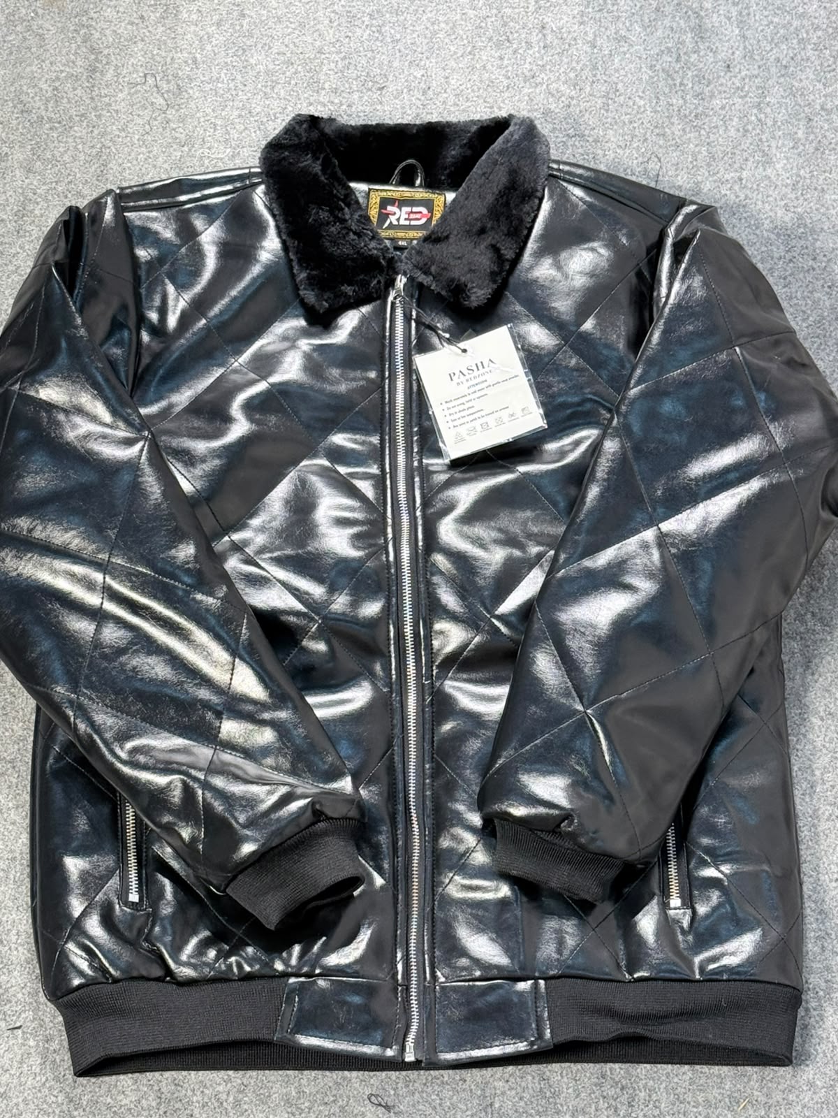 A black quilted faux-leather bomber jacket with a black faux-fur collar, shown flat on a gray textured background with a white retail tag attached.