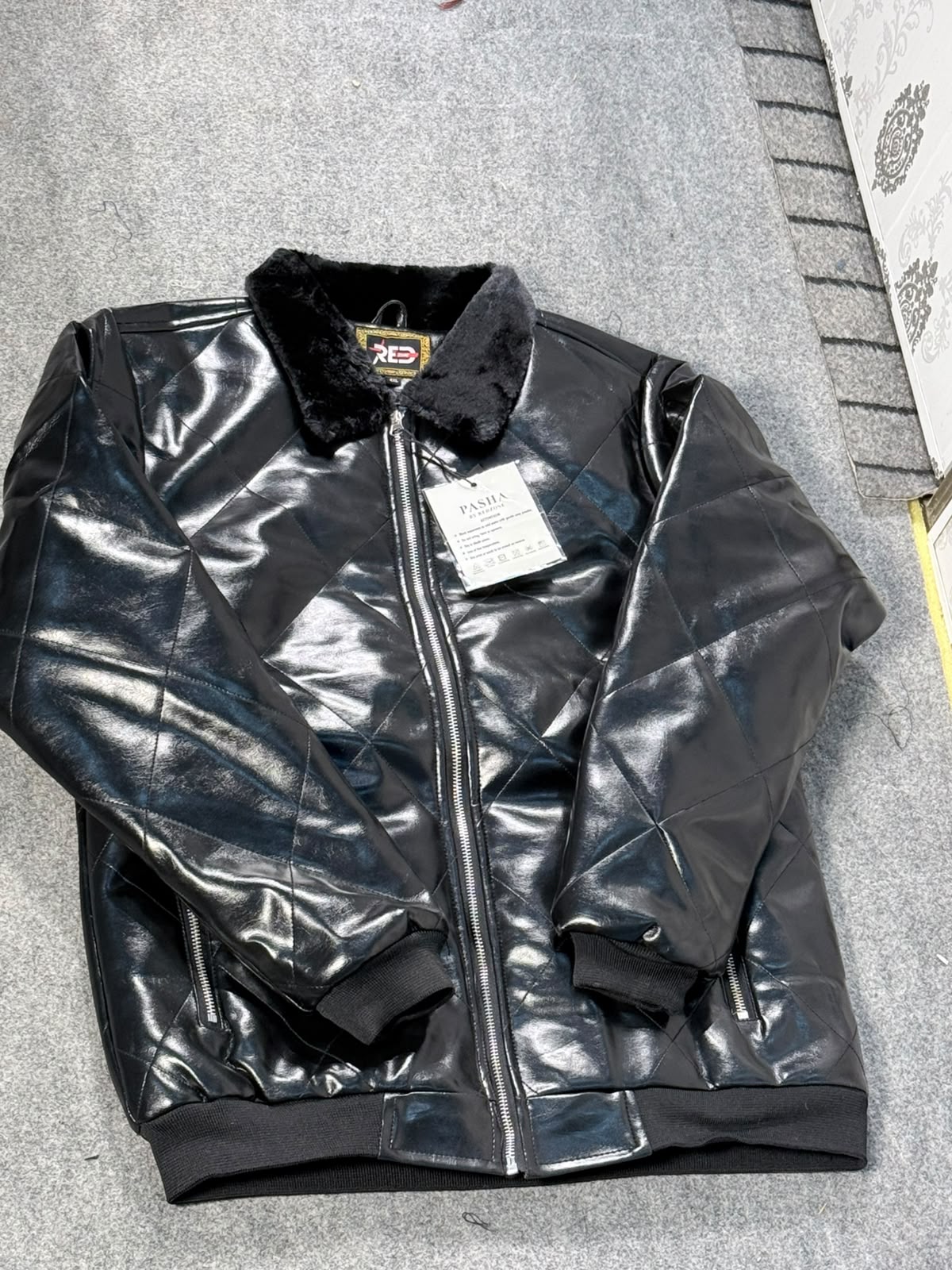 A black faux-leather bomber jacket with a fuzzy black collar and a front zipper, laid flat on a gray textured surface. A white retail tag is attached to the zipper.