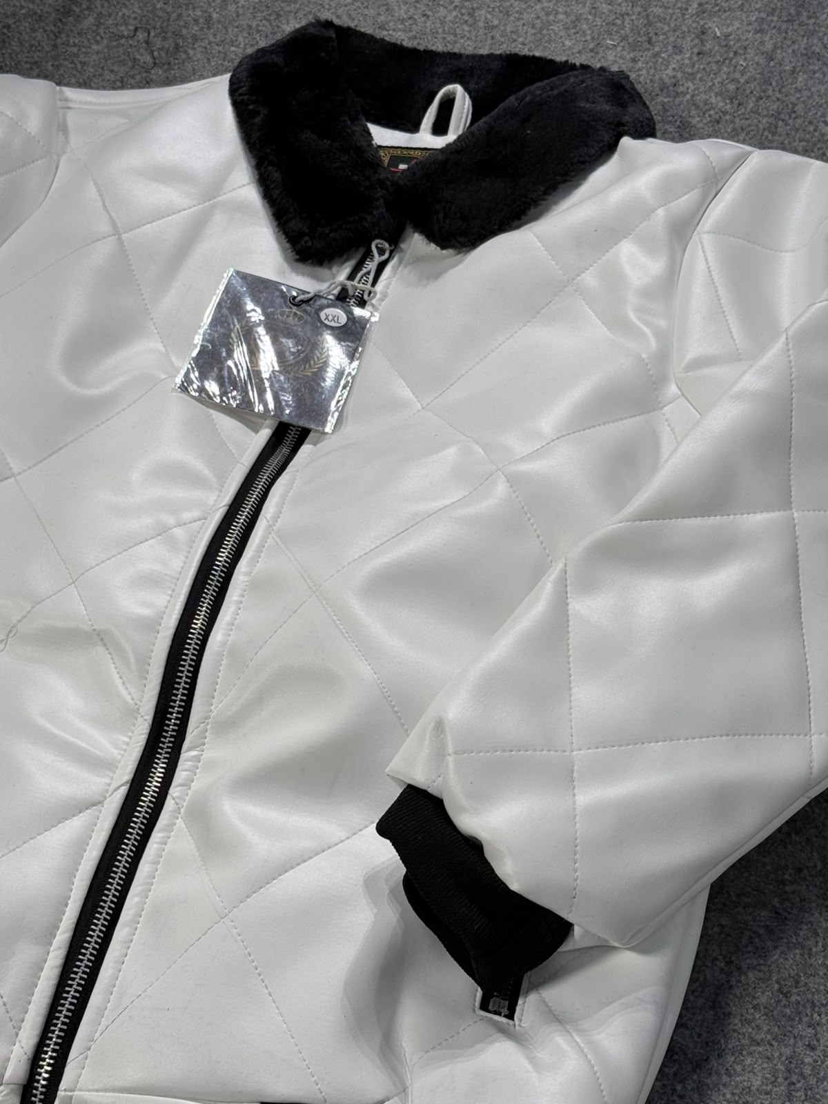 A close-up of a white quilted bomber jacket with a black faux-fur collar, black ribbed cuffs, and a black front zipper, shown with a silver hangtag on a gray textured background.