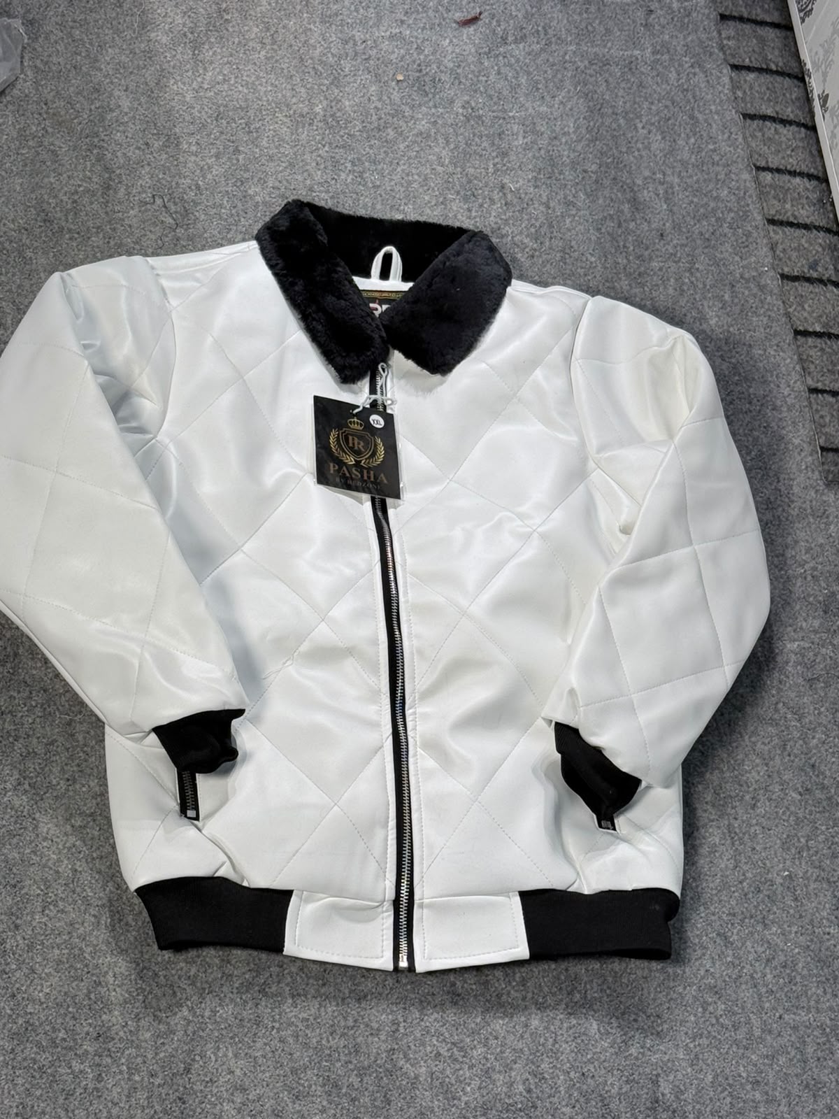 A white quilted bomber jacket with black ribbed trim, a black collar, and a black zipper, laid flat on a gray carpet. A hangtag is attached to the zipper.