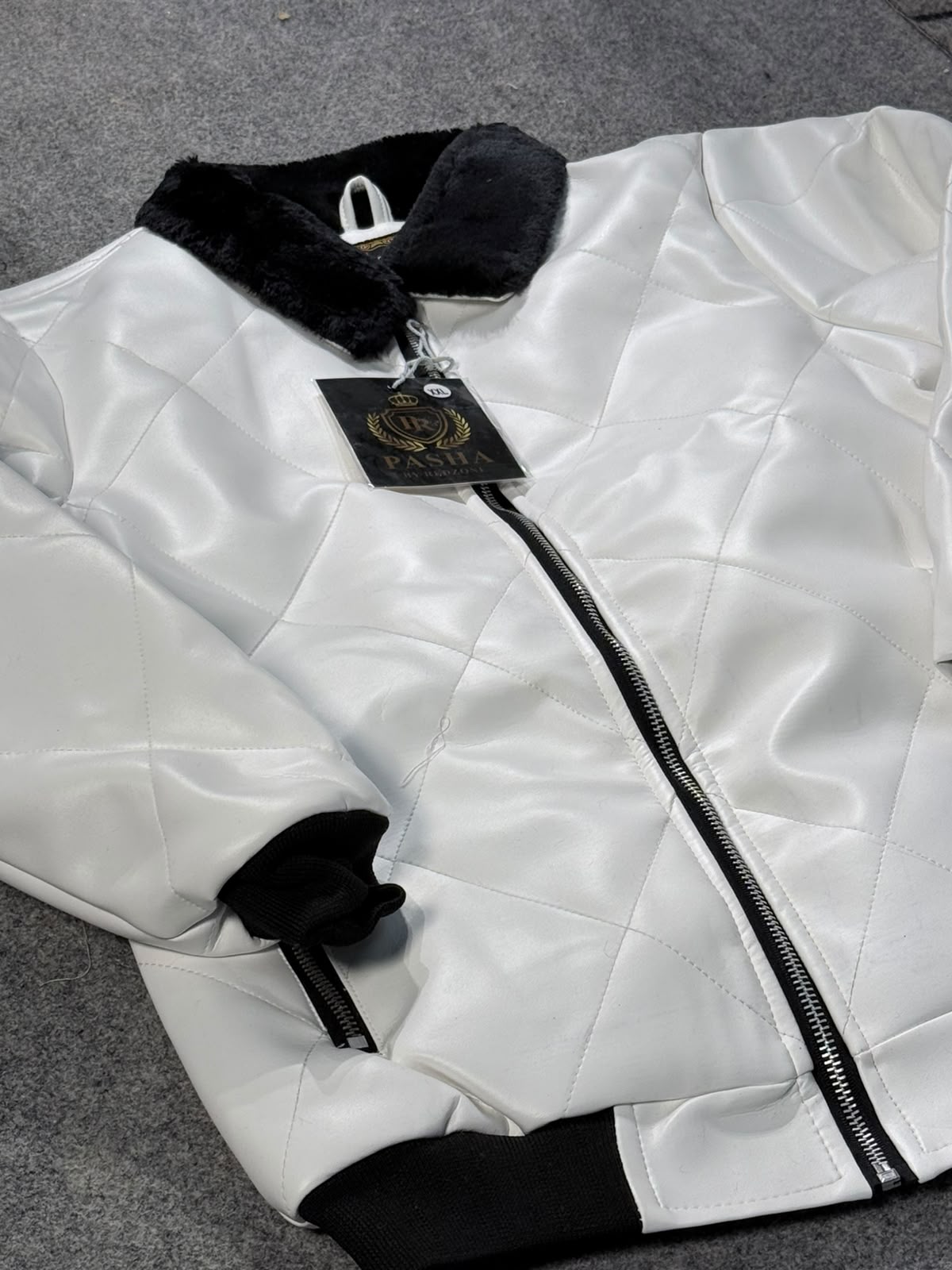 A white quilted faux-leather bomber jacket with black faux-fur collar, black ribbed cuffs, and a black zipper, shown with a hanging tag on a gray textured background.