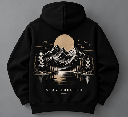 A black pullover hoodie shown from the back, featuring a large graphic print of a mountain landscape with a lake, pine trees, birds, and a large sun or moon. The text 'STAY FOCUSED' is printed below the graphic in white.