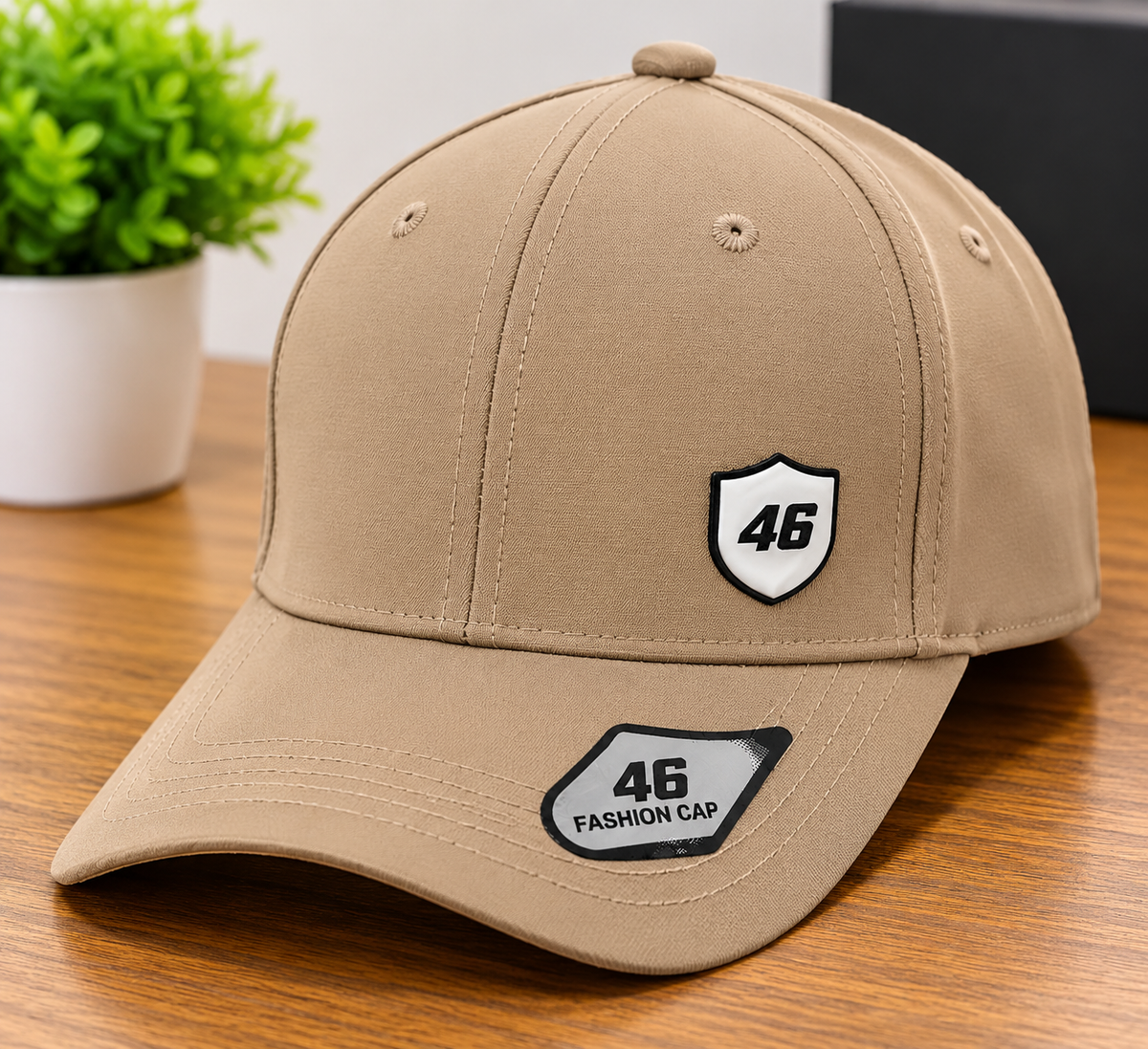 A tan baseball cap with a curved brim, featuring a white shield patch with the number 46 on the front and another patch on the brim, displayed on a wooden table with a small green plant in the background.