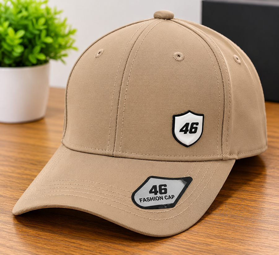 A tan baseball cap with a curved brim, featuring a white shield patch with the number 46 on the front and another patch on the brim, displayed on a wooden table with a small green plant in the background.