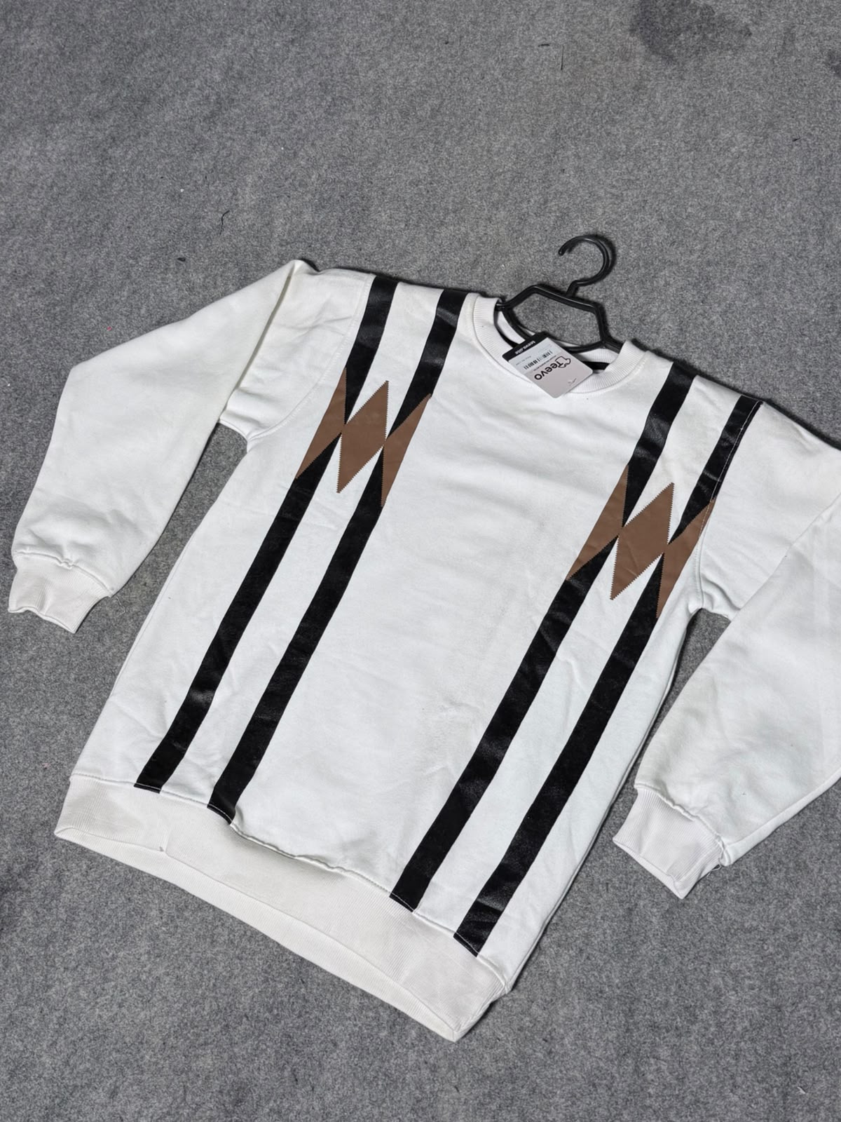 A white crewneck sweatshirt with bold black vertical stripes and brown lightning-bolt motifs on the chest, laid flat on a gray textured surface with a black hanger.