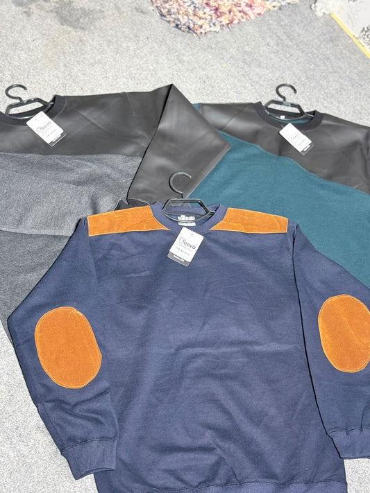 A navy blue crewneck sweatshirt with orange elbow patches and orange shoulder yoke, displayed on a hanger over other color variants (gray and teal) on a gray carpeted floor.